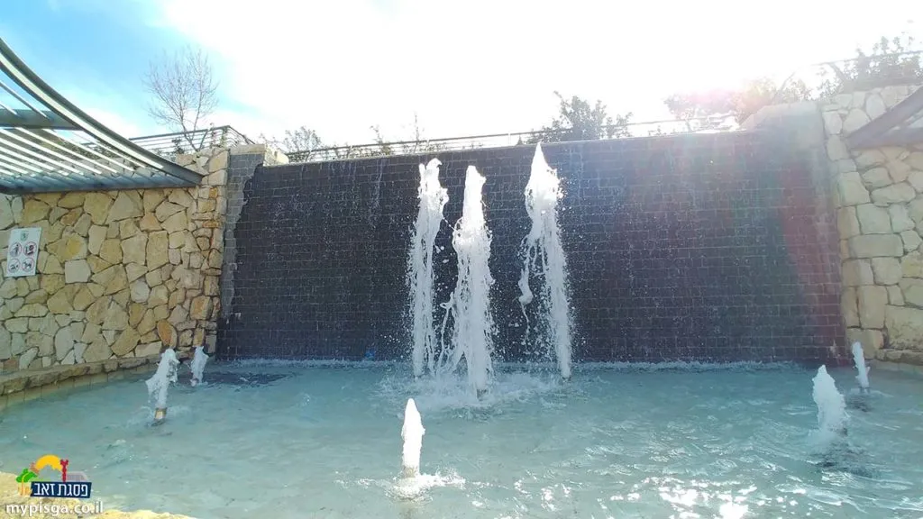 מפל מים על קיר אבן כהה בפארק עמנואל מורנו, ומזרקות שמתיזות סילוני מים בבריכה רדודה מוקפת אבן, ולידה פרגולה