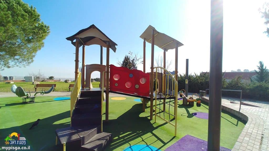 מתקן משחק עם מדרגות, סולם ומחיצה אדומה על משטח דשא סינטטי בפארק שמואל תמיר, ספסלים ונדנדת קפיץ ברקע ושמיים כחולים