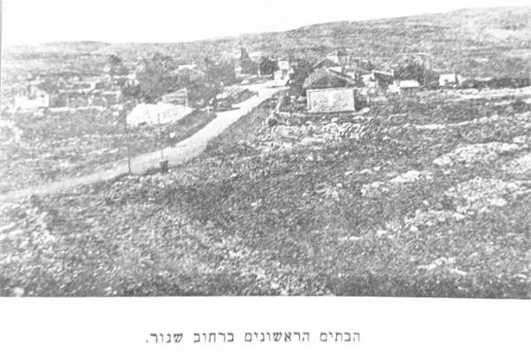 צילום שחור-לבן ישן: דרך עפר בהירה חוצה שטח סלעי וריק, ובצידה בתים קטנים בודדים, צריפים ועצים מעטים. מתחת לתמונה כיתוב בעברית: הבתים הראשונים ברחוב שנור