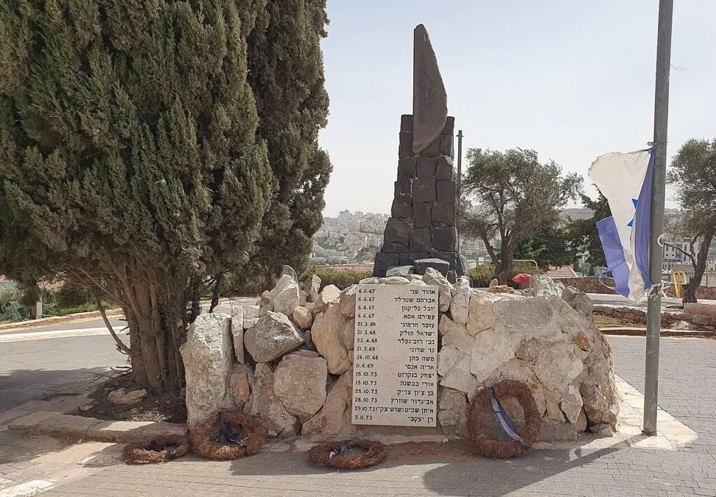 אנדרטת סיירת דוכיפת: עמוד אבן כהה וגבוה על ערימת אבנים לבנות, ובחזיתה לוח עם שמות חרוטים. לרגליה זרים, לצידה עץ ברוש ותורן עם דגל ישראל