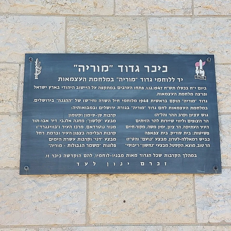 שלט מתכת כהה על קיר אבן בהירה. בראשו הכיתוב כיכר גדוד מוריה, יד ללוחמי גדוד מוריה במלחמת העצמאות, ומתחתיו שתי עמודות של שמות קרבות ומבצעים, ובשורה האחרונה: זכרם ינון לעד