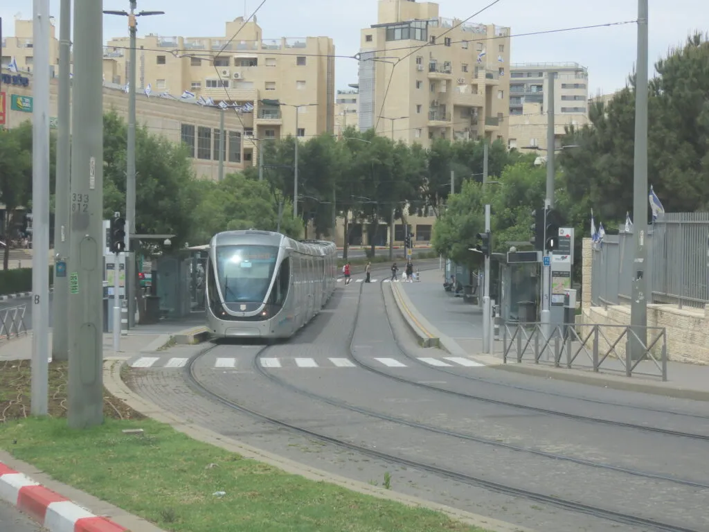 רכבת קלה כסופה נכנסת לתחנת פסגת זאב מרכז בשדרות משה דיין, משני צדי המסילה עצים ורציפים מקורים, וברקע בנייני אבן ובניין הקניון