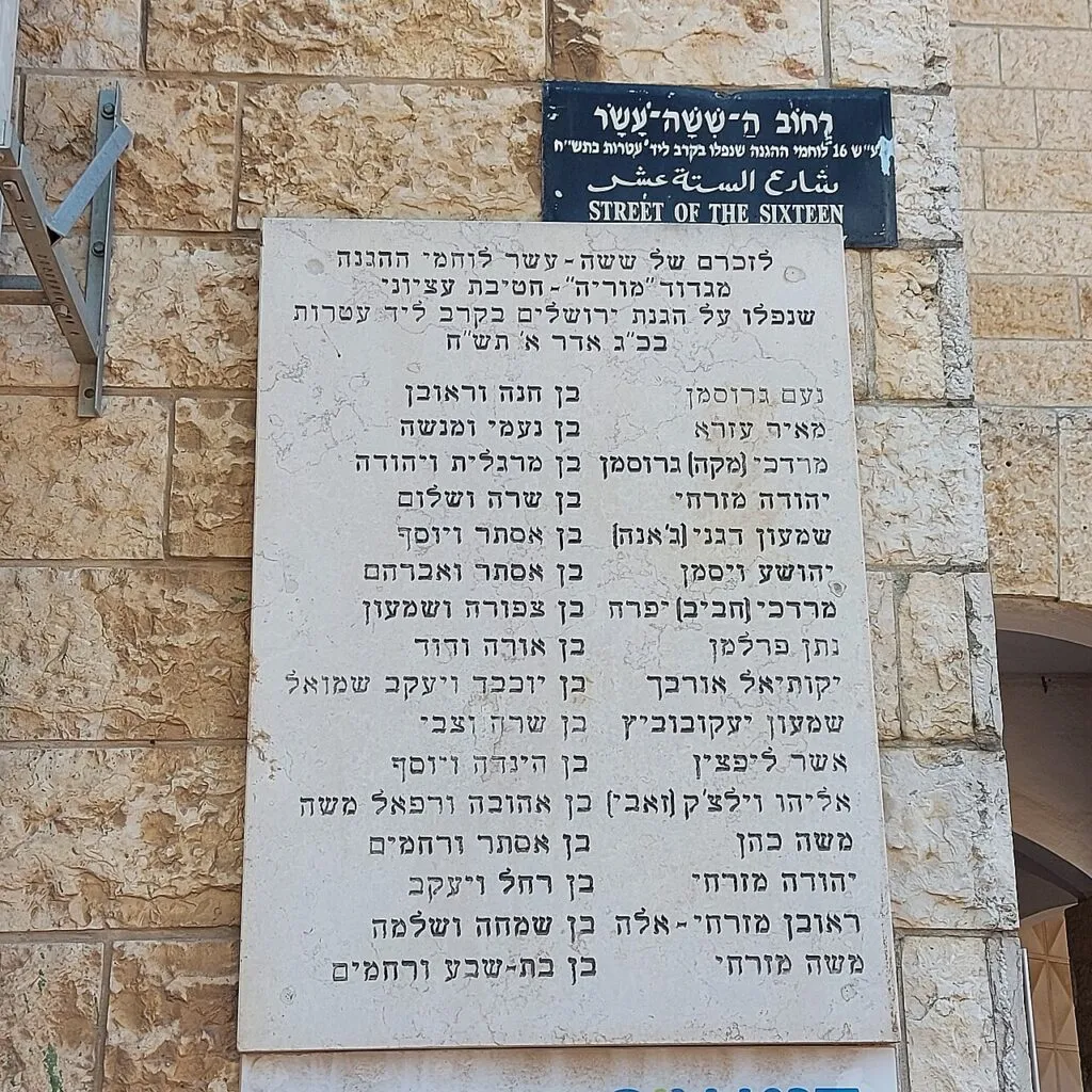לוח שיש לבן על קיר של אבן ירושלמית, ועליו חרוטים שמות שישה עשר לוחמי גדוד מוריה ולצד כל שם שמות ההורים. מעל הלוח שלט רחוב כחול כהה: רחוב השישה עשר, בעברית, בערבית ובאנגלית