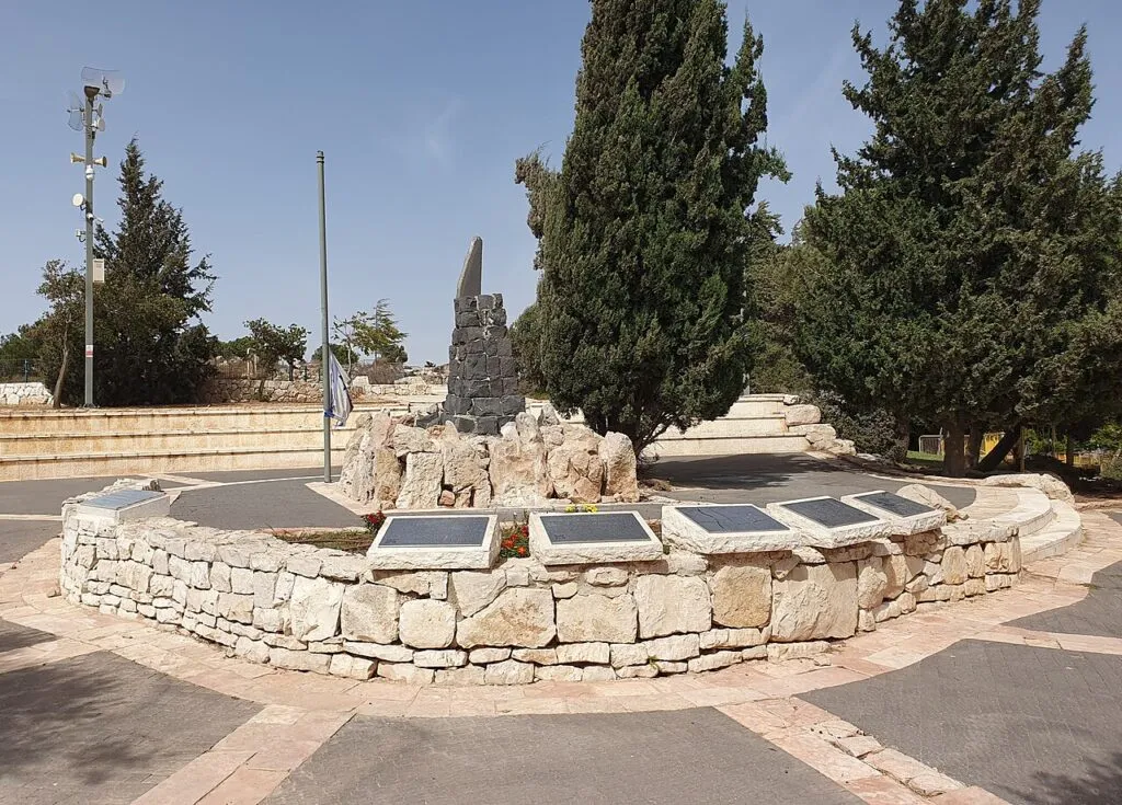 רחבת הזיכרון של סיירת דוכיפת: קיר אבן נמוך ומעוגל ועליו שורה של לוחות מתכת, ובמרכז עמוד אבן כהה וגבוה. מאחור עצי ברוש, עמוד תאורה ושמיים כחולים