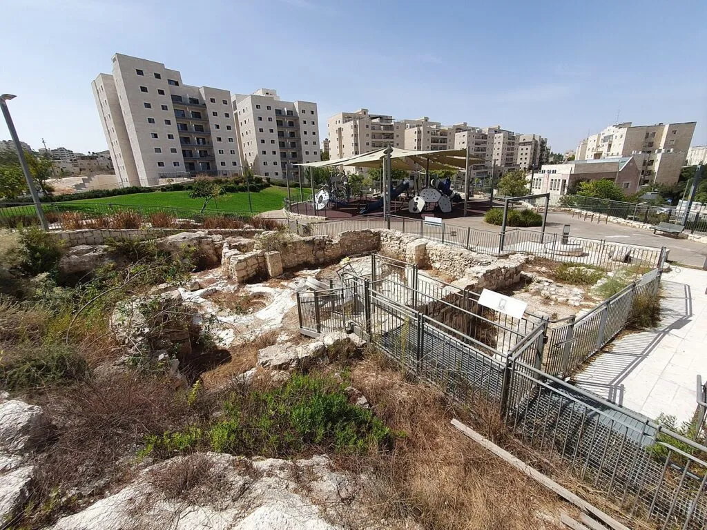 שרידי קירות אבן עתיקים מוקפים מעקה מתכת בחורבת עדסה, ומאחוריהם שביל מרוצף, מדשאה, גן משחקים מוצל ובניינים חדשים