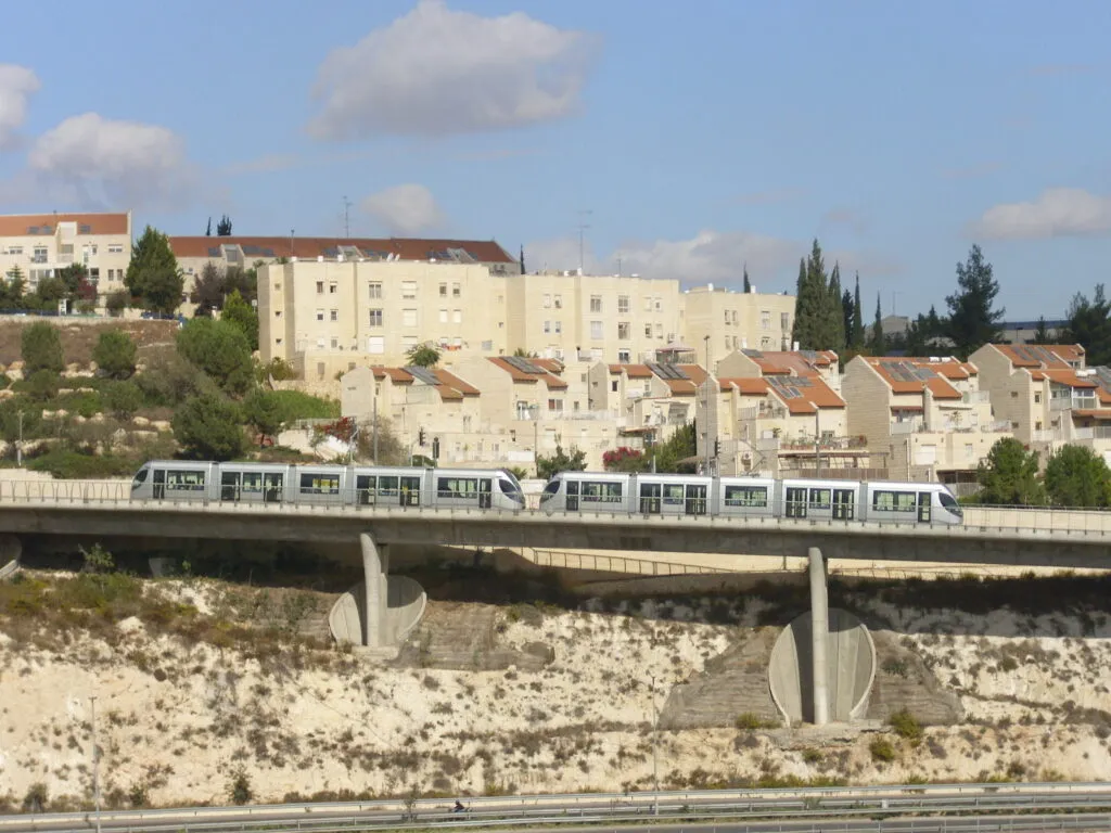 שתי רכבות קלות כסופות חוצות גשר בטון על עמודים, מתחת לגשר מדרון סלעי וכביש, וברקע בתי אבן עם גגות רעפים אדומים ועצים