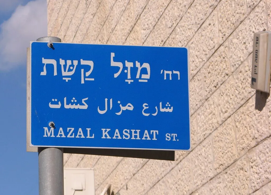 שלט רחוב כחול בעברית, בערבית ובאנגלית שעליו כתוב רחוב מזל קשת, תלוי על עמוד מול קיר אבן בהירה, ולידו שלט קטן של שדרות משה דיין