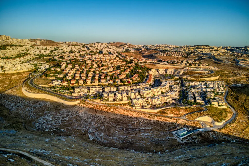מבט מהאוויר על שכונת מגורים על גבעה בשעת שקיעה: בתים פרטיים עם גגות רעפים אדומים, בנייני אבן בהירים נמוכים, כביש שמקיף את השכונה ומדרון סלעי בחזית