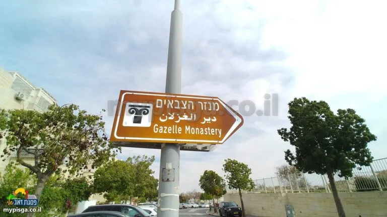 שלט חום עם הכיתוב 'מנזר הצבאים' וחץ, על עמוד ברחוב, עצים ומכוניות ברקע