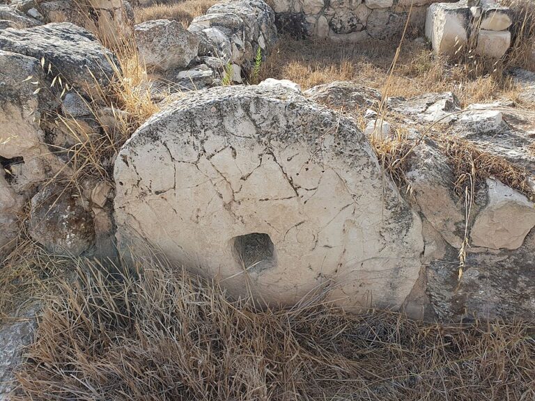 אבן עגולה וגדולה שעומדת על צידה, עם חור מרובע קטן, בין קירות אבן ועשב יבש