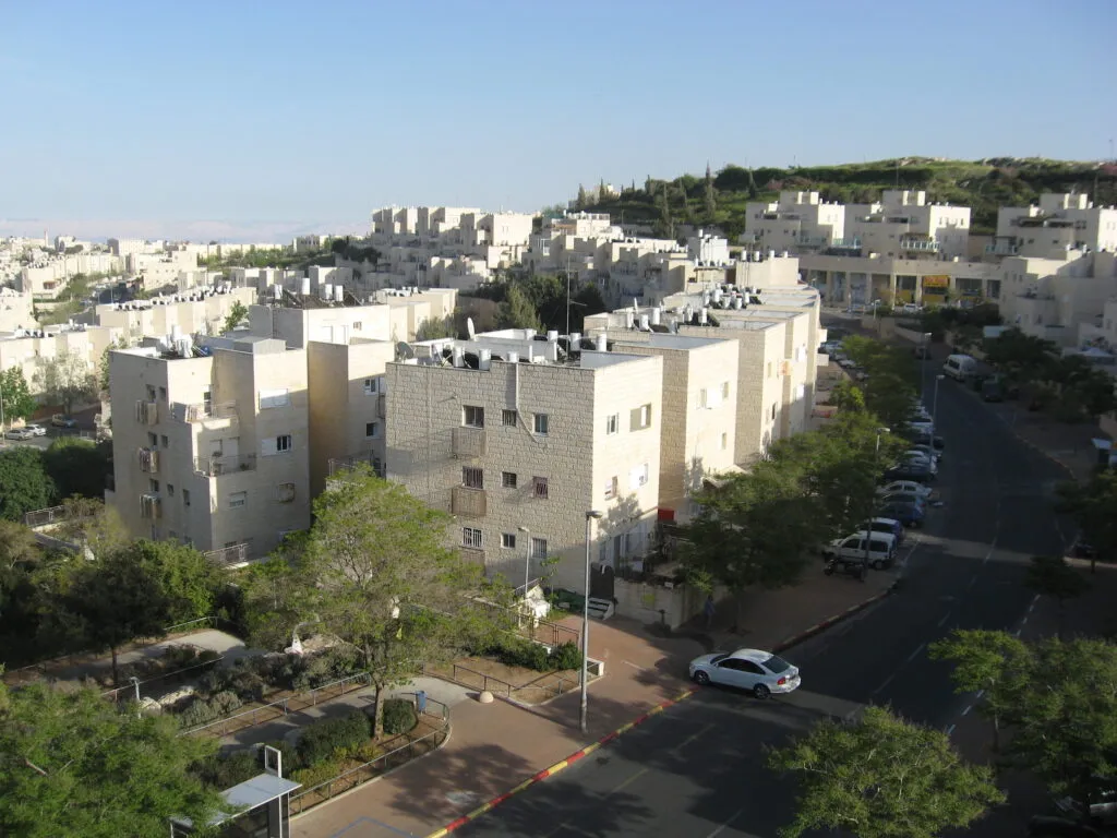 בנייני מגורים מאבן בהירה בני ארבע קומות לאורך רחוב עם עצים ומכוניות חונות, וברקע עוד בניינים וגבעה ירוקה