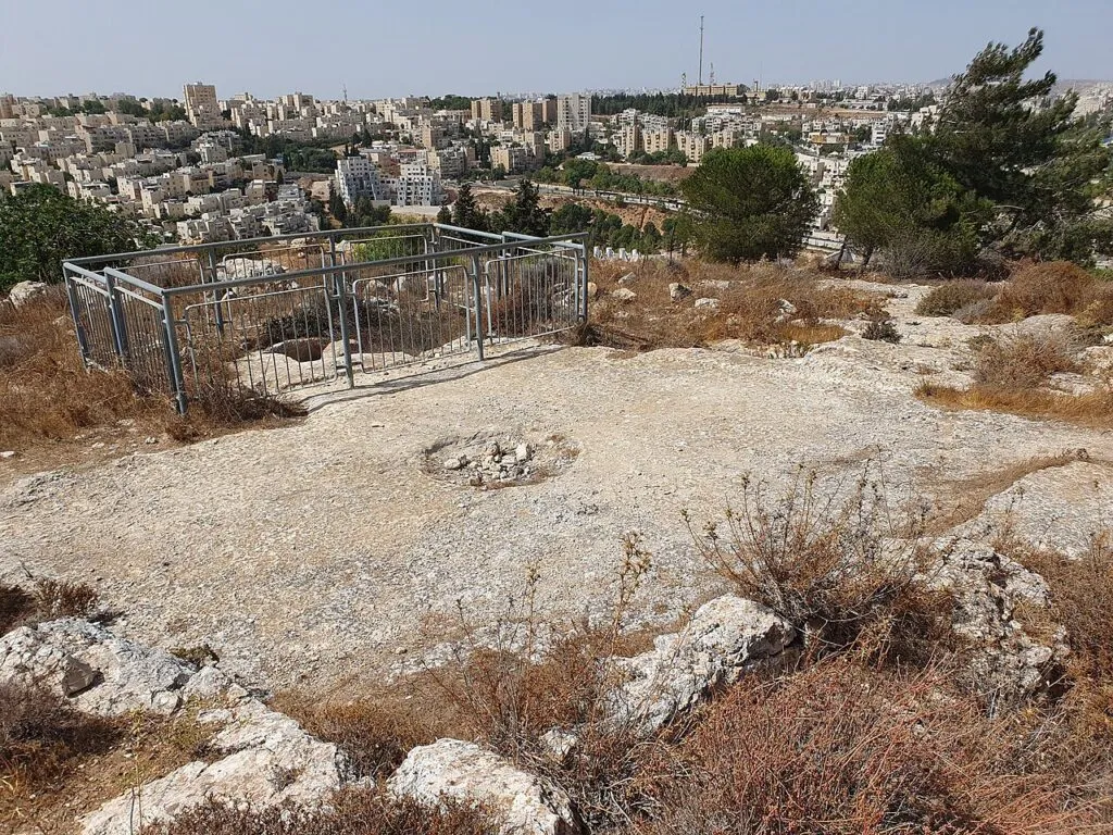 ראש הגבעה הארכיאולוגית בפסגת זאב מרכז: משטח סלע עם חציבות, גדר מתכת סביב בור עתיק, ובניינים של ירושלים ברקע